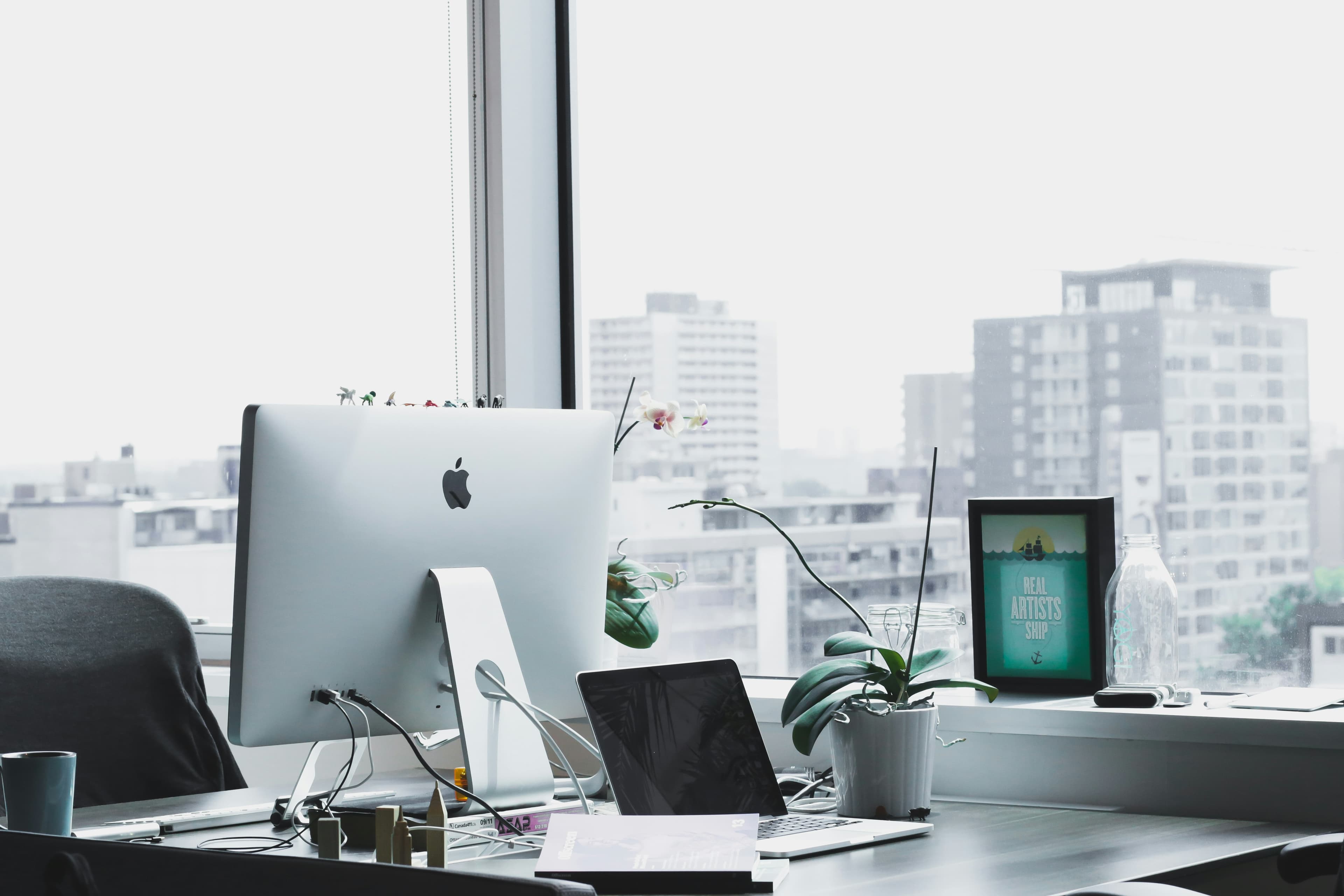 A quiet desk with a desktop monitor, a small plant and a notebook in front of a window.