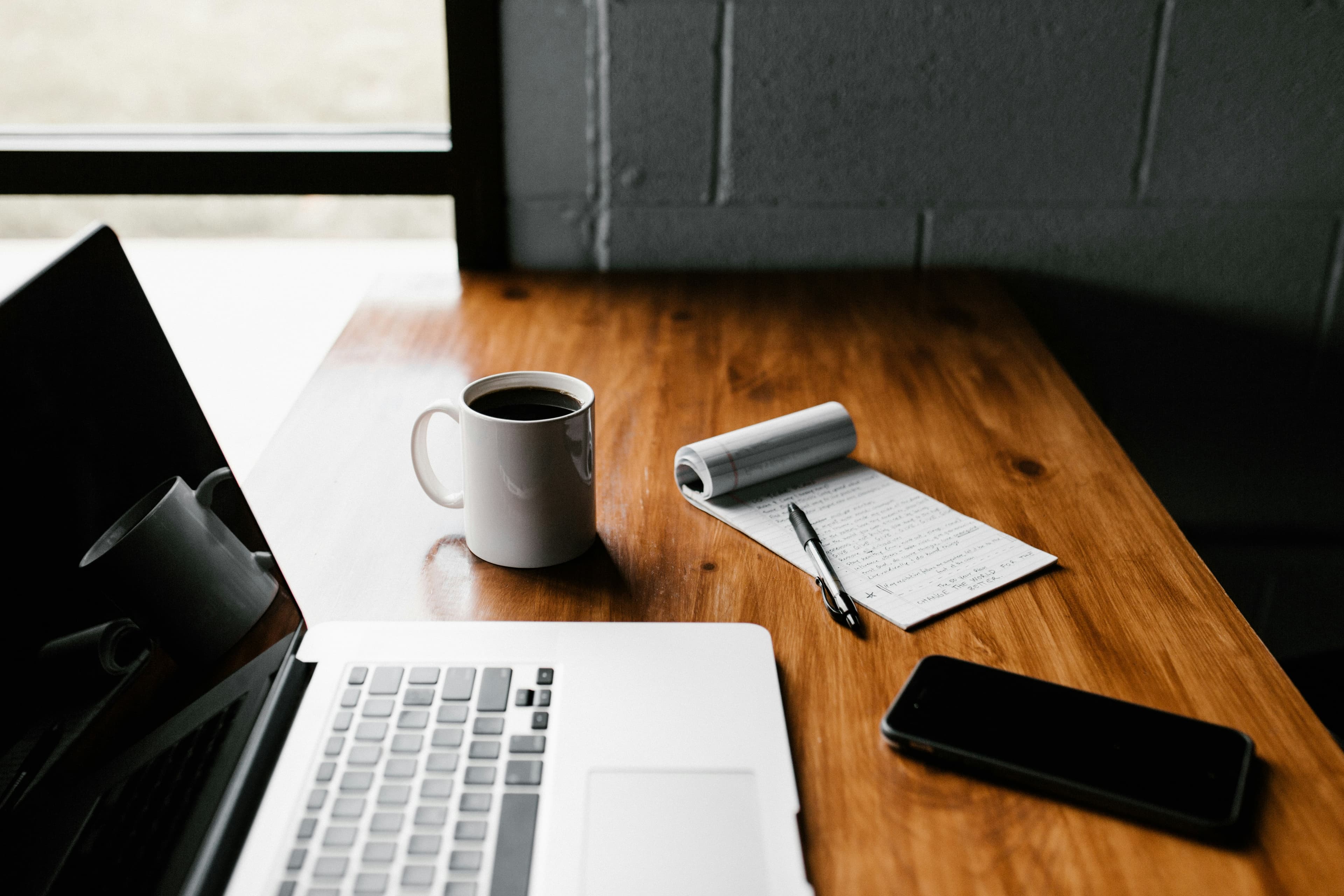 A laptop open on a wooden desk next to a cup of coffee, a phone and a handwritten notepad.