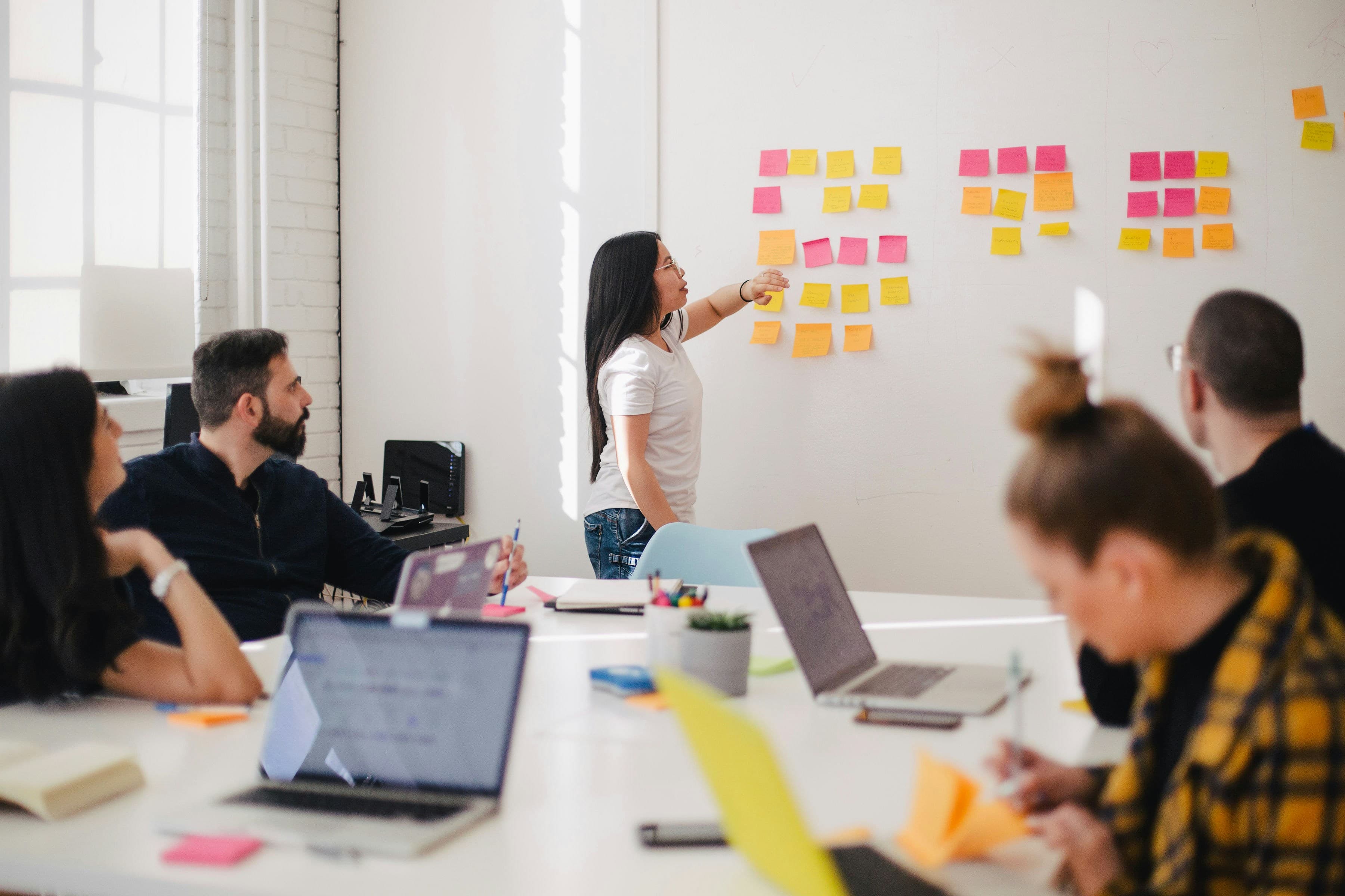 A team meeting where one person arranges sticky notes on a wall while colleagues work at a table.